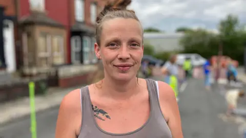 A white woman wearing a grey vest top is smiling at the camera. She is wearing her blonde hair up in a bun. Behind her children are playing in a residential street that has been blocked off to traffic. 