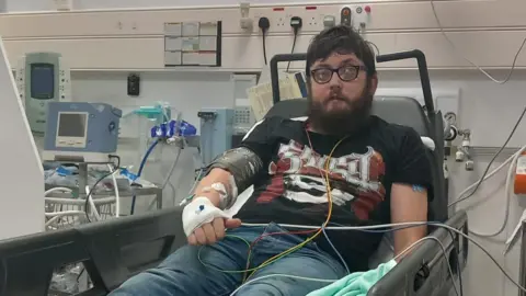 Jake Waine A man in a hospital bed with man tubes attached to him.