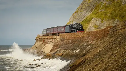 Martin Price A train pulled by a steam engine runs along a track between a crashing wave in the sea and very steep cliff.