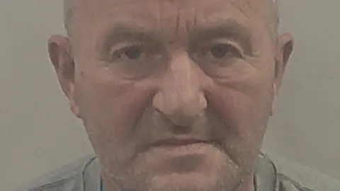 Close-up custody shot of Michael. He has thin receding grey hair and stubbly beard, and is wearing a grey top.