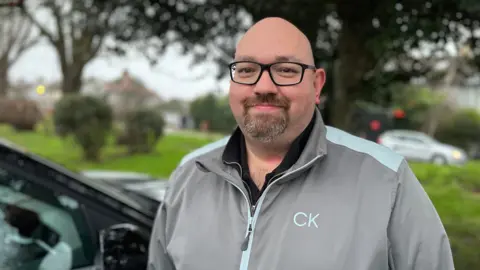 The image shows a driving instructor named Dan Carter. He is smiling at the camera. He has black square-shaped glasses on. He has a small beard. He is wearing a grey jacket which has light-blue panelling on the shoulders. The zip lining is also the same shade of blue. Underneath he has a black quarter-zip on. Behind him is grass and trees. To the bottom left of the picture is a black car. 
