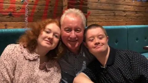 Mark Jones Mark and his children sit together at a restaurant table with drinks and cutlery in front of them. Behind them is a wooden wall with colourful writing, and a birthday balloon featuring a cartoon character is visible to the left. 