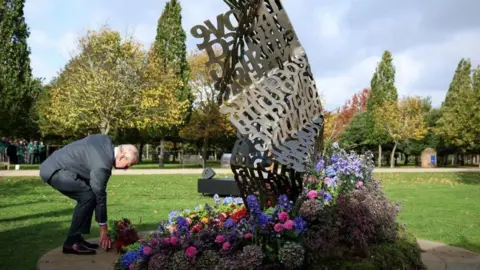 Reuters The King stoops to lay flowers at the foot of a twisted metal sculpture made up of lots of individual words and phrases, shaped like a large scrunched piece of paper. It is a sunny day at the National Memorial Arboretum and the trees are bathed in sunlight in the background.