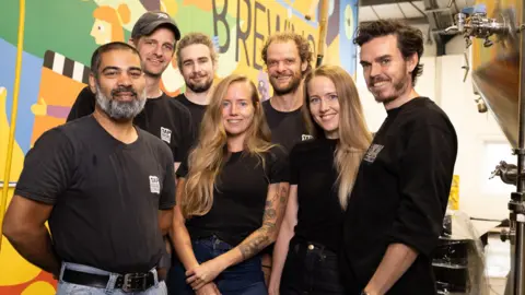 Tap Social The Tap Social Movement team smiling for a picture at the brewery, including founders Tess and Amy Taylor, and Paul Humpherson, who are in the first row.