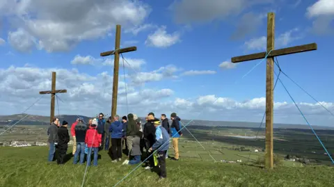 Three wood crosses on a green hill, each attached to the ground with blue rope. There is a group of people, around 20, gathered in front of the middle cross.