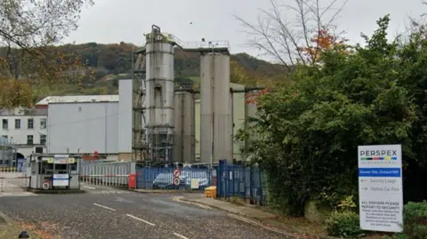 Street view of the site in Darwen showing tall cylindrical industrial towers behind a blue fence and a gate with security barriers. There are bushes and a "Perspex" sign to the right