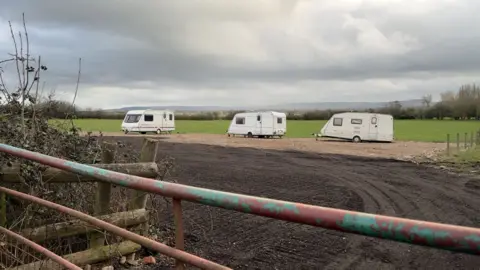 Three caravans parked in a field which has had a hard standing laid down. A metal gate can be seen in the foreground and there are hills in the distance.