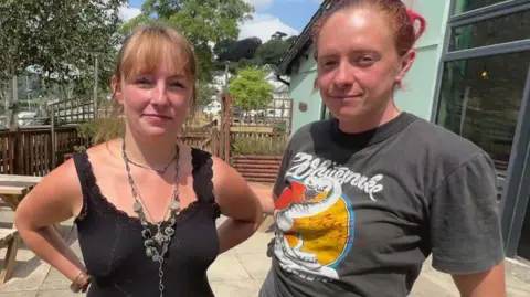 Kerenza is wearing a black vest top and a chunky necklace. Ellie is wearing a grey tshirt with a Whitesnake logo. Both women have their hair tied back. They are standing in a patio which has a wooden bench.