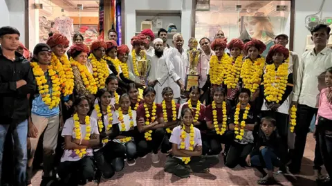 A girls' football team are pictured in sports clothing, wearing yellow pom-poms. Two men standing among them are holding trophies