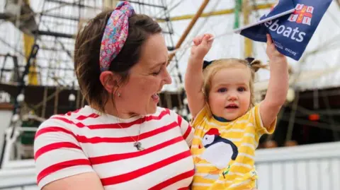 Woman in white and red striped top and pink headband carrying toddler daughter who is holding an RNLI flag in the air. They are in front of a tall ship.