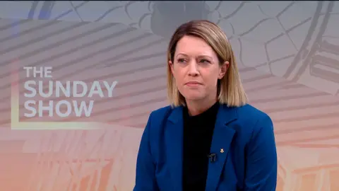 A woman with short blonde hair and wearing a blue suit, sits in a TV studio, the backdrop shows branding for The Sunday Show