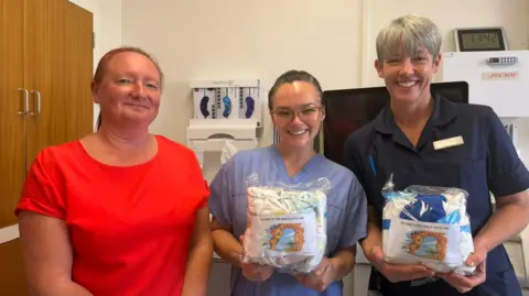 Three women stand in a line in a hospital delivery suite. The woman on the left has a red T-shirt on and her auburn hair is tied back. The other two women are nurses and are wearing blue uniforms. They are holding packages containing clothes and nappies for new babies.
