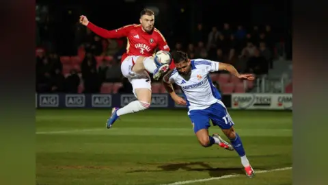 Two footballers on the pitch, both mid-air: the Salford player is almost fully at the height of his opponent's shoulder while he aims to kick the ball