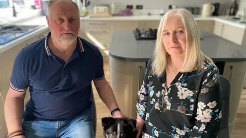 Christine and Neil Clayton, a middle-aged man and woman, sit in their kitchen with serious expressions on their faces. They each have a hand on a dog which is sitting between them. 