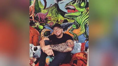 Adam Salisbury Artist Adam Salisbury wearing a black baseball cap, black t-shirt and jeans, sits on his son's bed with his painting of dinosaurs he created on the bedroom wall. He is smiling. 