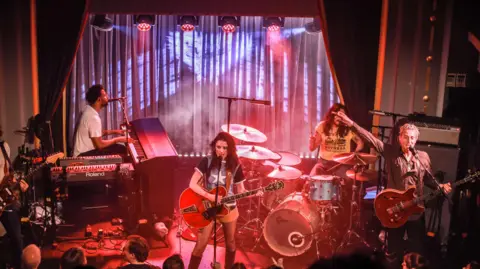 Debbie Hickey/Getty Images Wolf Alice performs on stage in front of a curtain, with a singer playing guitar, and other members playing keyboards, bass guitar and drums. The audience watches from below in dim lighting. 