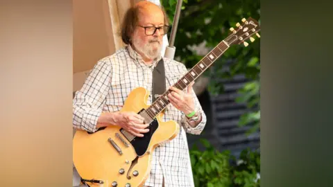 Mike Sandford A man plays guitar. He is in a white shirt and has glasses and a big grey beard. 
