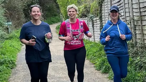 Alice Draper Three women, two wearing blue, and one in a bright pink running t-shirt, running down a path with grass and bushes on one side and a fence on the other side. They are all smiling.