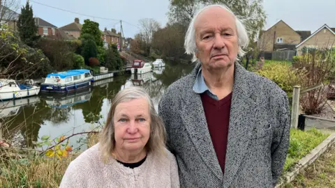 John Devine/BBC Tony Gosling, 75, on the right has grey hair and is wearing a herringbone black and white overcoat, with a purple jumper over a blue shirt, his wife, Patsy, 71, has grey and sandy coloured shoulder length hair parted to the left with a beige woollen fleece over a dark shirt. Behind them is a river with 4 cruiser boats moored on the far side, residential housing can be seen too.