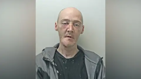 Peter Vernal, a man wearing a grey jacket over a black top with white marks on it, stares vacantly forward in a mugshot.