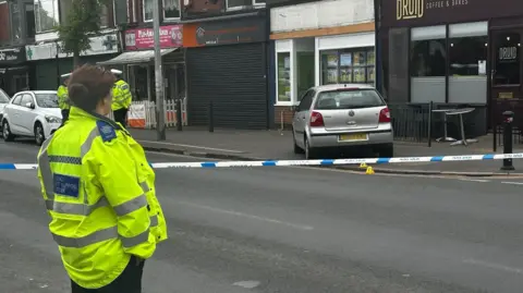 BBC A silver car has mounted the pavement outside a cafe. There is a blue and white police cordon around the scene. There is an police officer wearing a yellow hi-visibility jacket and black trousers, with her brown hair tied in a bun, standing with her back to the camera to the left of the scene. There are two police officers in the same uniform, also wearing hats, standing talking in the distance.