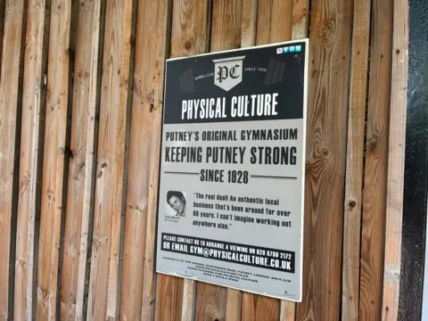 Charlotte Lillywhite/LDRS Black, white and grey sign that says "Physical Culture. Putney's original gymnasium keeping Putney strong since 1928." It includes a quote and the gym's contact details at the bottom. The sign is screwed into a background into a wooden wall. 