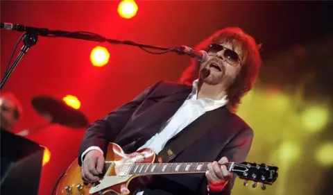 Jeff Lynne plays a guitar and sings into a microphone on stage. He wears a purple suit jacket and a white shirt