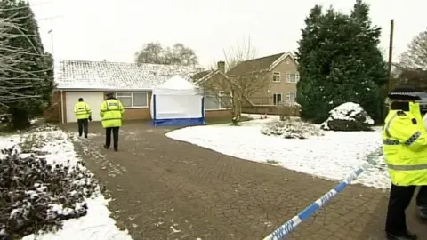 A snow-covered bungalow and snow-covered garden. Four police officers, wearing yellow coats, can be seen in the bungalow driveway. The end of the driveway is cordoned off with blue and white tape marked with the word "police". A white forensics tent can be seen directly outside the house.