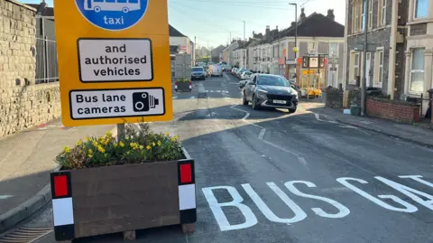 A newly-installed Bus Gate on Barton Hill. There is a plant pot with a sign signalling the bus gate on the left of the road and white paint on the street saying Bus Gate.