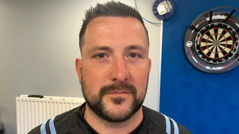 BBC Shane McDonald has short black hair pushed back from his forehead and a dark beard. He is wearing a sports top and a dart board is on the wall on the right of the picture.