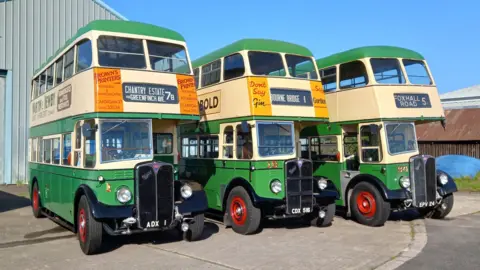 Ipswich Transport Museum The three classic double decker buses are lined up outside a barn. The sky is blue and the buses are painted green and white, while the wheels are bright red.