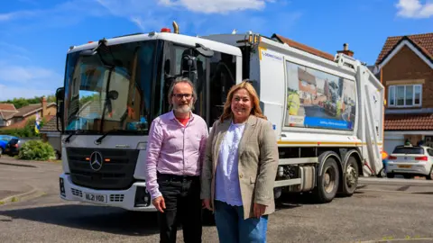 Steve Harker (left) and Lisa Evans standing in front of a bin wagon in Ingleby Barwick. Mr Harker has long hair and a grey beard. He is wearing glasses and a red shirt and dark jeans. Ms Evans has long blonde hair and is wearing a white top and beige blazer. The bin lorry is white. Houses can be seen in the background.
