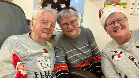 Piers Hopkirk/BBC An elderly man, elderly woman and younger woman all wearing Christmas jumpers and hats smiling in a room 