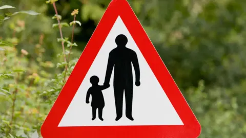 A red and white triangular road sign with a grown-up holding a child's hand 
