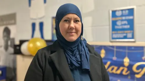 Shariqua Ahmed/BBC Laura, a woman wearing a blue headscarf and a green coat, smiling for the camera.