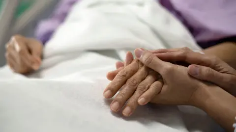 Patient's hand being held by a relative while they lie on a hospice bed