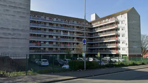 Google A block of flats, with several storeys high, with a lamp post outside, and cars parked outside the building. 