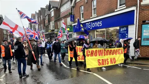 Dozens of protestors on a high street behind a banner that reads 'Crowborough Says No'. Many are carrying UK or England flags.