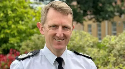 West Mercia Police A man with short brown hair, wearing a white shirt with black tie and epaulettes is standing smiling at the camera. There is a building behind some bushes in the background.