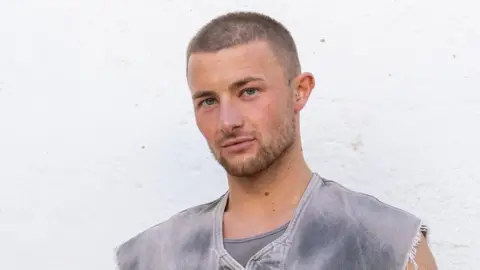 Jordan Burrow looking into the camera with pursed lips. He is standing in front of a white wall. He has shaved brown hair and is wearing a grey top with cut-off sleeves.