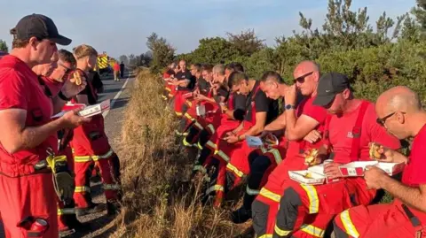 Firefighters in red uniform, eating burgers, while standing on the roadside.