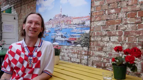 Ed sits at a yellow table in an outdoor courtyard wearing a red and white Croatia football shirt, with a brick wall, potted plants and a large photograph of a coastal town behind him.