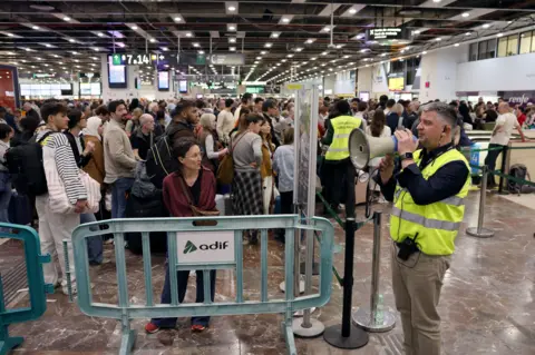 EPA Crowds at Barcelona Sants railway station wait for updates on 28 April, 2025, as a man in a high-viz jacket stands to the right of the picture facing left and speaking into a megaphone. A woman stands centre-left in the foreground facing the camera and looking left. In front of her is a crowd barrier.