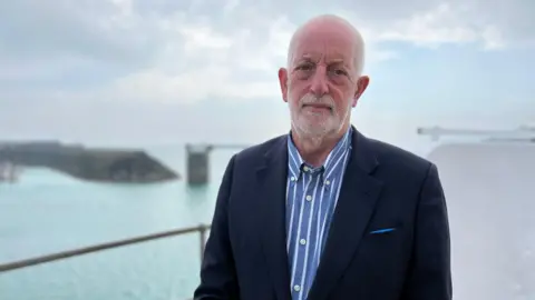 Steve Luce is smiling at the camera. He has short white hair and a beard and is wearing a striped shirt and blazer. To his left is a bay and a quay.