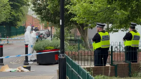 BBC/Chloe Laversuch The image shows a cordoned-off residential street with trees, parked cars, and a small green space bordered by railings. Two uniformed police officers stand on the right side of the image behind a low green metal fence. Both officers are wearing high-visibility yellow vests. A black lamppost stands near the centre of the frame. Blue-and-white police tape stretches across the road. Two individuals wearing white forensic suits with gloves are positioned on the left side of the street.
