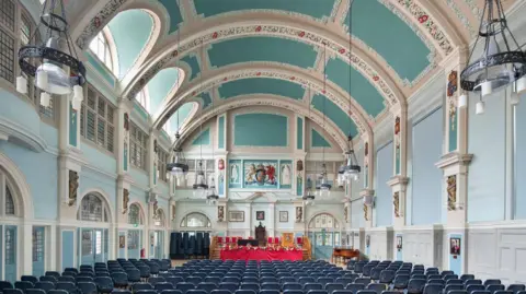 A central hall with pale blue and green walls and ceiling with blue chairs and ornate plasterwork 