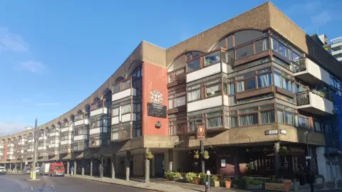 A large, modern building with a curved facade. The structure is multi-story, featuring wide windows and balconies on each level. Its exterior combines brick and concrete, with some areas painted red. Above an entrance, signage reads “Corporation of London" below the City of London Corporation crest. The sky is clear and blue.