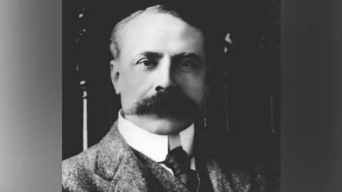 A black and white, head and shoulders shot of Edward Elgar, with a large moustache and wearing a white shirt and tie with a suit jacket