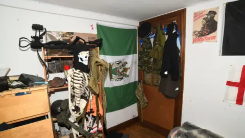PA Media A view of the corner of the accused's bedroom. Military clothing is hanging on the back of the door, a poster with a historic Nazi image on it hangs on the wall and a large weapon and plastic skeleton wearing a mask are in the foreground. There is also a green and white flag hung sideways between the door and the skeleton.