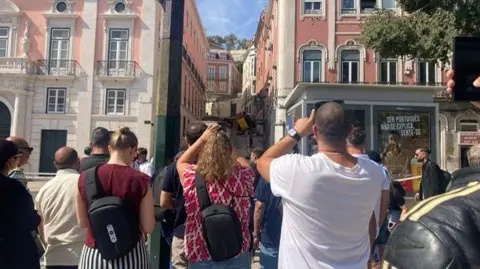 Several people take photos at the bottom of a hill in Lisbon of the funicular crash.